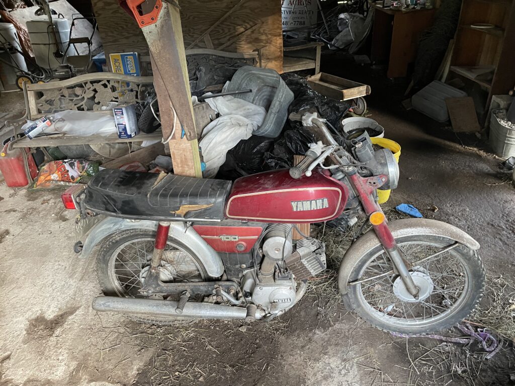 Image of a red motorcycle in a messy barn, covered in dust. 